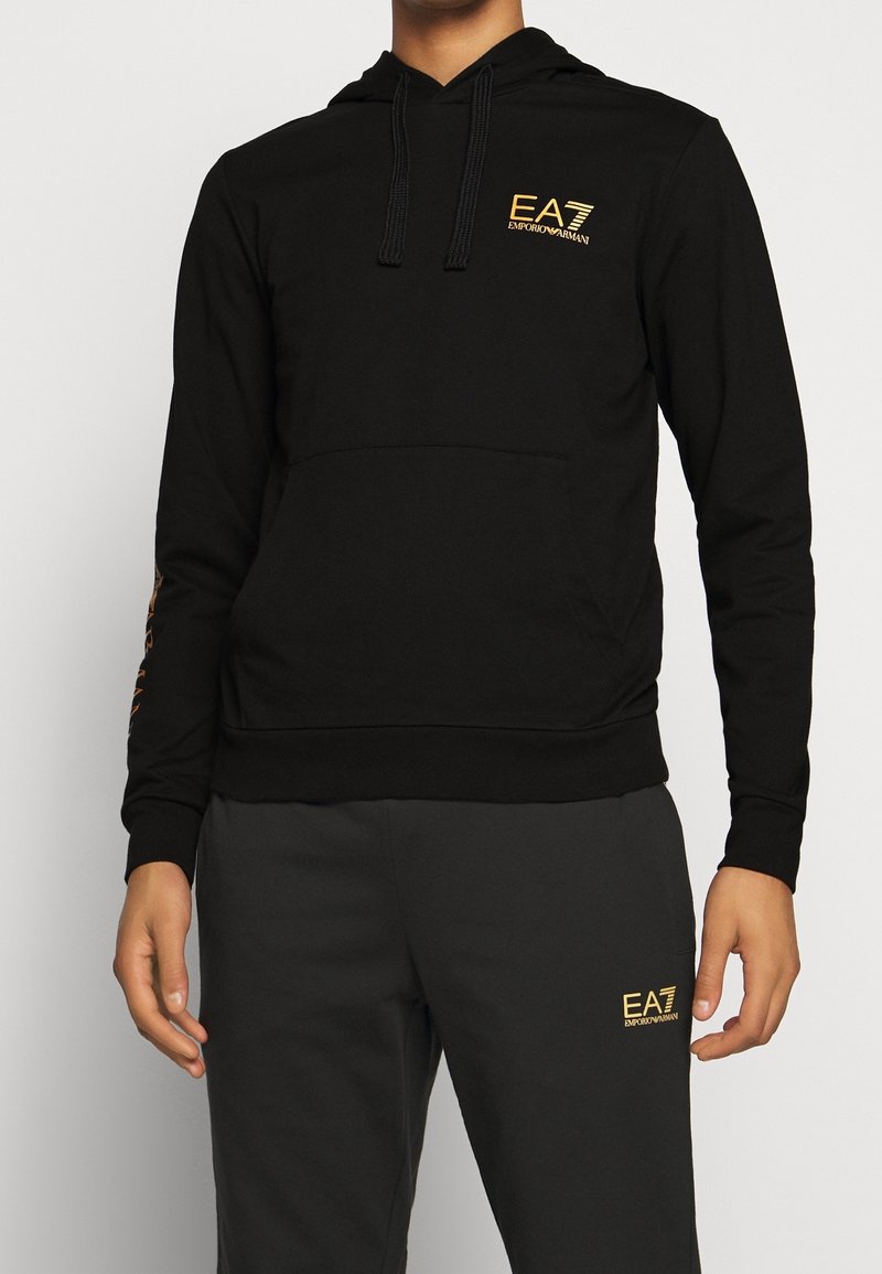 Černá mikina s klokankou, která má na hrudi zlaté logo EA7 a dekorativní design na rukávech. Měkký bavlněný materiál.