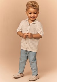 Lys beige strikket skjorte med knapper og korte ærmer, kombineret med lyseblå bukser og beige loafers, stående mod en neutral baggrund.