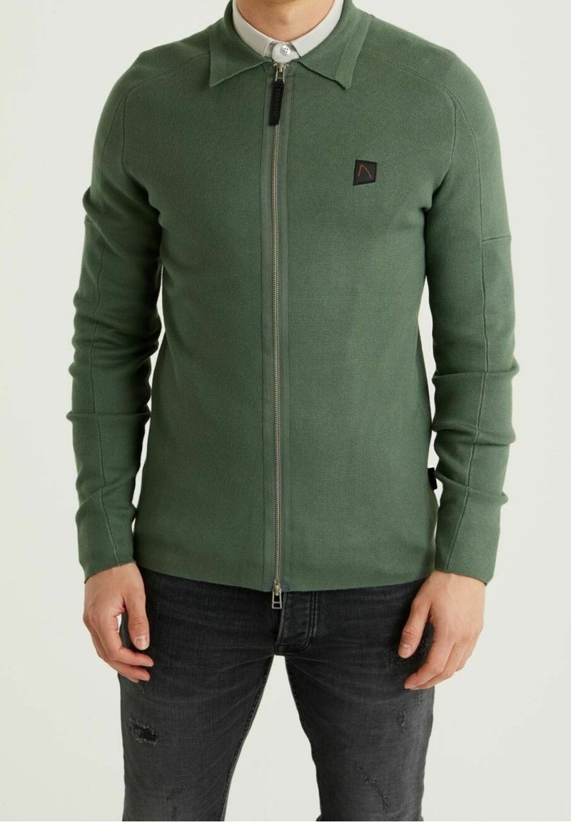 Homme portant une veste en maille verte zippée sur une chemise gris clair à col, associée à un jean noir déchiré, debout les mains détendues.