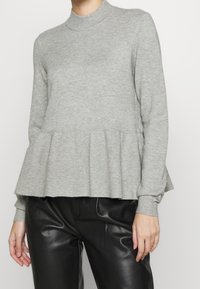 Kobieta w jasnoszarym swetrze z długim rękawem i peplum, zestawionym z czarnymi spodniami z ekologicznej skóry.