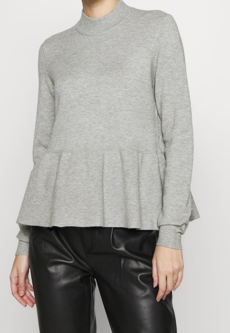 Kobieta w jasnoszarym swetrze z długim rękawem i peplum, zestawionym z czarnymi spodniami z ekologicznej skóry.