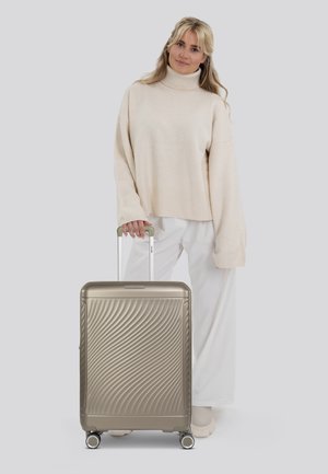 Valigia rigida dorata con texture ondulata, manico estensibile e quattro ruote, abbinata a un maglione color crema e pantaloni bianchi.