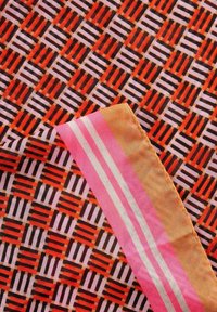 Multikleurige stof met een geometrisch patroon van rode, roze en zwarte rechthoeken. Roze en witte gestreepte rand aan één kant. Zachte textuur.