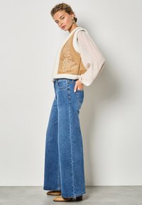 Tamarisierter bestickter Weste mit weißem Kunstfellbesatz, kombiniert mit weit geschnittenen blauen Jeans und braunen Absatzstiefeln vor einem neutralen Hintergrund.