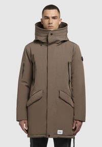 Jachetă parka călduroasă, de culoare maro, cu glugă și guler înalt, cu buzunare cu fermoar și clapă, din țesătură texturată și șnururi de reglare. Pată logo notabilă.