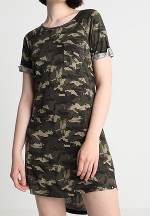 Femme portant une robe à manches courtes imprimée camouflage avec des revers aux manches et une poche poitrine, debout devant un fond uni.