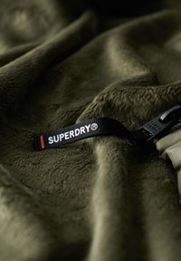 Olive zielony miękki materiał o puszystej fakturze; na powierzchni materiału widoczny jest czarny pasek z logo "SUPERDRY®" oraz detal z klamrą.
