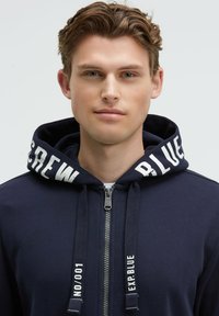 Sudadera con cremallera de color marino, con grandes letras blancas en la capucha y en los cordones. Tejido suave con un diseño simple y cierre frontal de cremallera.