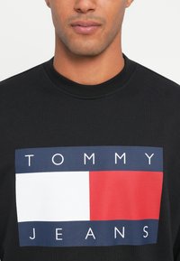 Hombre con un sudadera negra de cuello redondo con un logo rectangular de Tommy Jeans en azul, blanco y rojo en el pecho.