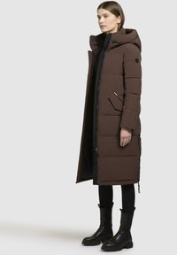 Cappotto puffer marrone con cappuccio, dotato di chiusura con zip e bottoni, tasche laterali e design alla lunghezza del ginocchio. Indossato sopra un abbigliamento nero.