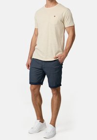 Camisa de manga corta beige claro con un bolsillo y un botón marrón, combinada con pantalones cortos oscuros de patrón azul y zapatillas blancas.