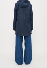 Osoba v tmavě modrém pláštěnce s kapucí a širokých modrých džínách stojící zády ke kameře na jednobarevném bílém pozadí.