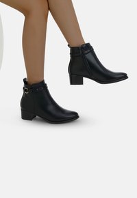 Stivali in pelle nera alla caviglia con tacco basso a blocco, cerniera laterale e decorativa indossati su gambe marrone chiaro su uno sfondo bianco.