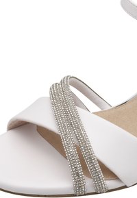Sandale blanche avec des brides entrecroisées, l'une ornée de strass argentés. La semelle intérieure est beige et légèrement texturée pour offrir une bonne traction.