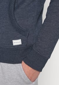 Felpa blu navy in morbido tessuto con tasca a marsupio, polsini a coste e una piccola etichetta bianca Tom Tailor di lato.