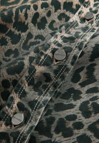 Mønstret stof med et grønt og beige leopardprint, med sølvknapper som lukning, struktureret overflade og synlige sydetaljer.
