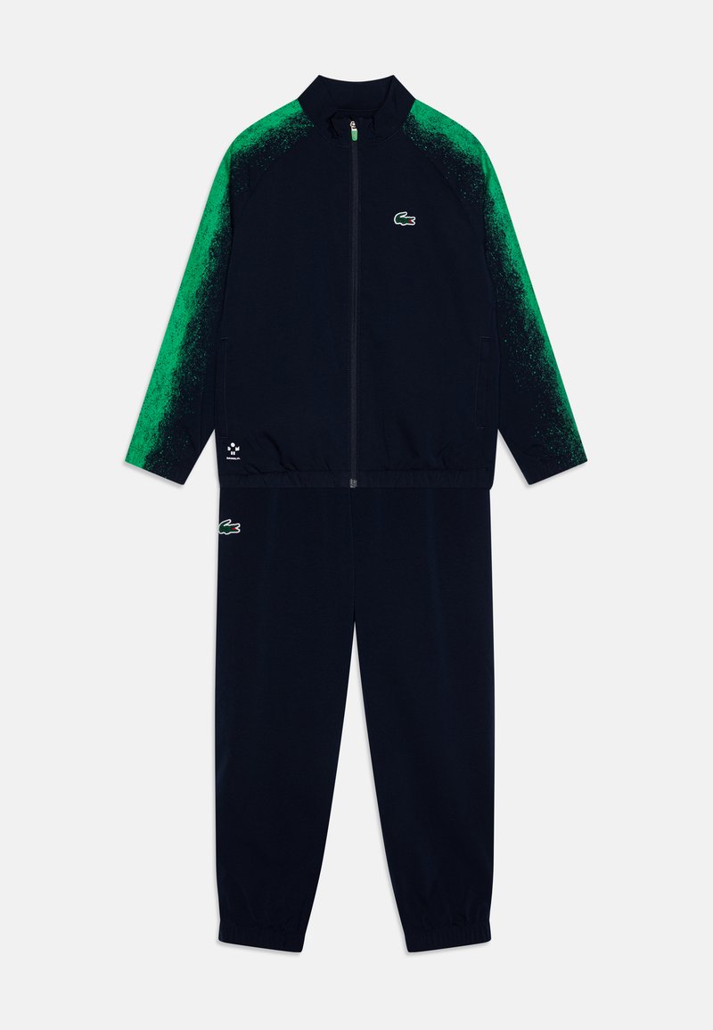 Survêtement marine avec des accents en dégradé de vert, veste zippée et pantalon élastiqué. Présente le logo Lacoste en haut à gauche et en bas à droite de la jambe.