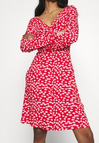 Czerwona sukienka z długimi rękawami, dekoltem w kształcie litery V i białym kwiatowym wzorem. Kształt w linii A, miękka tkanina i długość do kolan. Wzór zawiera małe stokrotki.