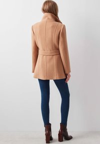 Lichtbruine wollen jas met een hoge kraag, ceintuur in de taille en verticale naaddetails, gecombineerd met blauwe skinny jeans en bruine haklaarzen.