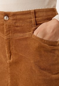 Pantalon en velours côtelé marron à coupe droite, avec une poche avant et une fermeture à bouton. Le tissu texturé présente de fines côtes verticales.
