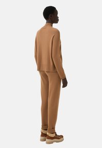 Tan knitted sweater with a high neck, paired with matching tapered pants. The outfit features ribbed cuffs and textured fabric. Brown lace-up boots.