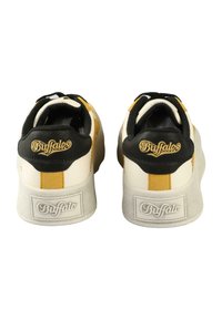 Lave sneakers med hvitt lærmateriale, svarte og gule aksenter, teksturert gummisåle og gylden logo på hælen.