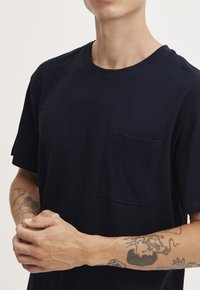 Tricou din bumbac de culoare bleumarin, cu mâneci scurte, decolteu rotund și un buzunar pe piept; țesătura pare netedă și ușor texturată.