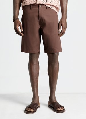 Homme portant un short marron jusqu'aux genoux, une chemise rose à motifs et des sandales marron tissées à enfiler, debout devant un fond blanc.