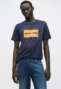 Μπλε βαμβακερό t-shirt με στρογγυλή λαιμόκοψη και κοντά μανίκια. Φέρει πορτοκαλί ορθογώνιο γραφικό με το κείμενο "JACK & JONES" σε μαύρο.