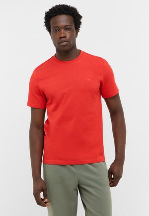 Red cotton t-shirt with short sleeves and a crew neckline, featuring minimal branding on the chest. Paired with light green pants.
