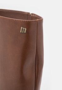 Tige de botte en cuir marron avec coutures visibles et petit logo métallique "m" près du bord supérieur sur un fond uni.