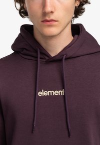 Sudadera morada con un logo frontal en beige claro, con capucha ajustable y textura suave. Sin patrones ni acentos adicionales visibles.