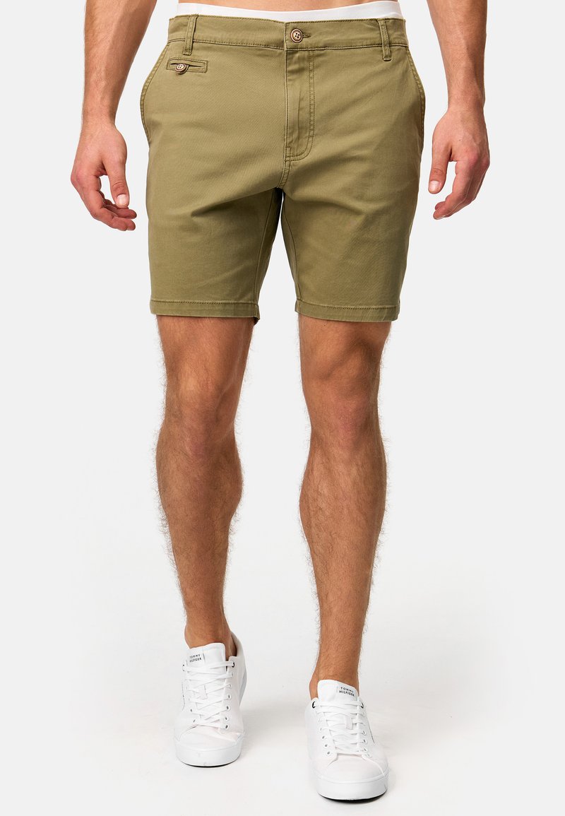 Olivgrüne Shorts aus Baumwolle mit Knopfverschluss, Seitentaschen und gerader Passform. Kombiniert mit weißen Sneakers.