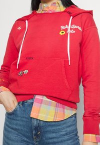 Červená bavlněná mikina s bílými šňůrkami, vyšívanými květinovými detaily a přední kapsou, vrstvená přes vícebarevnou kostkovanou košili.