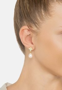 Gold earrings feature a floral design with a pearl drop. The floral part has sparkling accents, creating a contrasting texture.