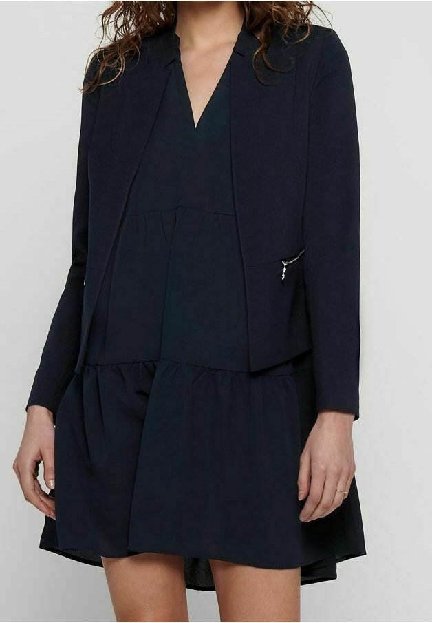 Blazer marine sur une robe noire, avec un décolleté en V, jupe évasée et poche latérale avec détail de fermeture éclair métallique. Tissu lisse.