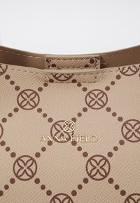 Sac à main beige texturé avec un motif circulaire marron et un logo doré "ANNA FIELD" centré sous une bordure en cuir.