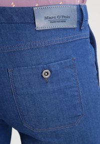Close-up of blue denim pants back pocket with button and stitched Marc O'Polo brand label on waistband.