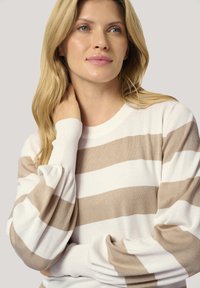 Pull en tricot beige et blanc à rayures avec un col rond à côtes et des manches bouffantes, offrant une texture douce et une coupe décontractée.