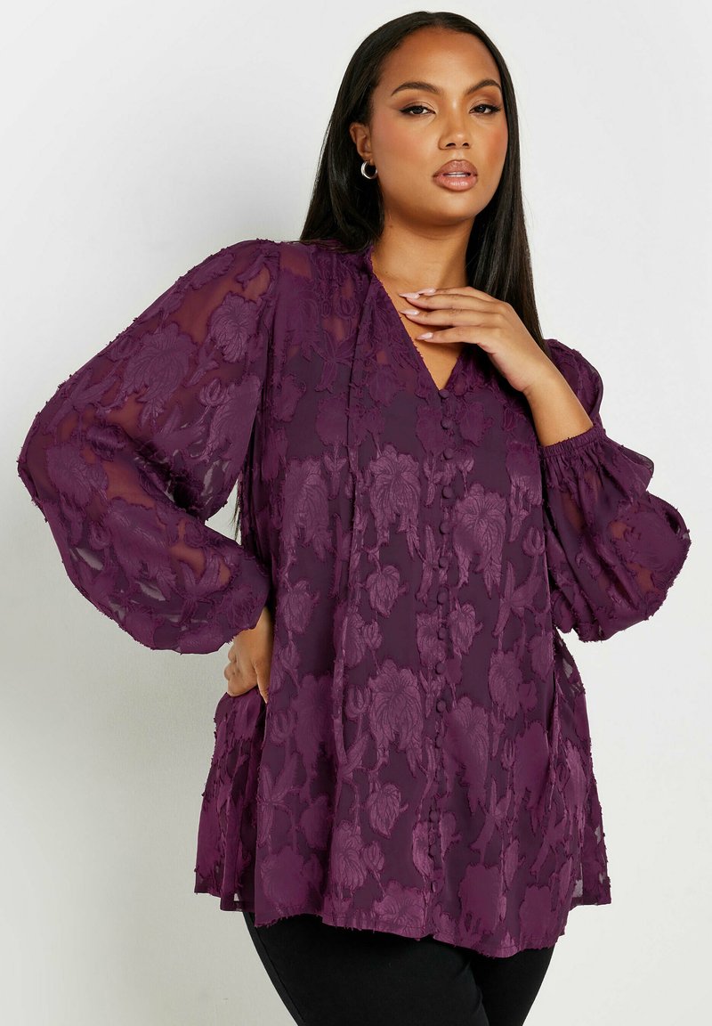 Blusa viola con ricami floreali, scollo a V e maniche lunghe a sbuffo. Presenta dettagli con bottoni e una vestibilità rilassata, sovrapposta a una base uniforme.