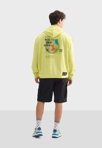 Felpa gialla con un design grafico sul retro, abbinata a pantaloni corti neri e sneakers multicolori. Include un cappuccio e polsini a coste.