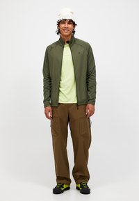 Giacca verde oliva con zip sopra una maglietta verde chiaro, abbinata a pantaloni cargo marroni con molteplici tasche e sneaker nere con accenti fluorescenti.