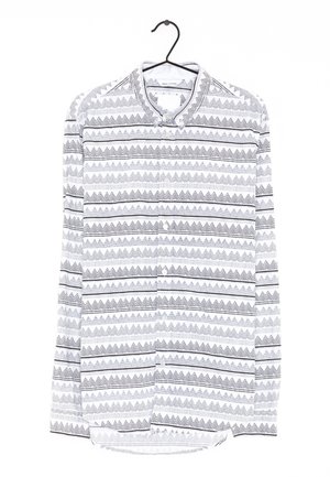 Biała koszula na długi rękaw z czarnymi geometrycznymi wzorami zygzaków i trójkątów, zawieszona na czarnym wieszaku na białym tle.