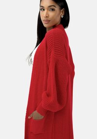 Cárdigan rojo de punto con manga larga, de corte holgado y dos bolsillos frontales. Tejido texturizado con un diseño de punto grueso y abierto.
