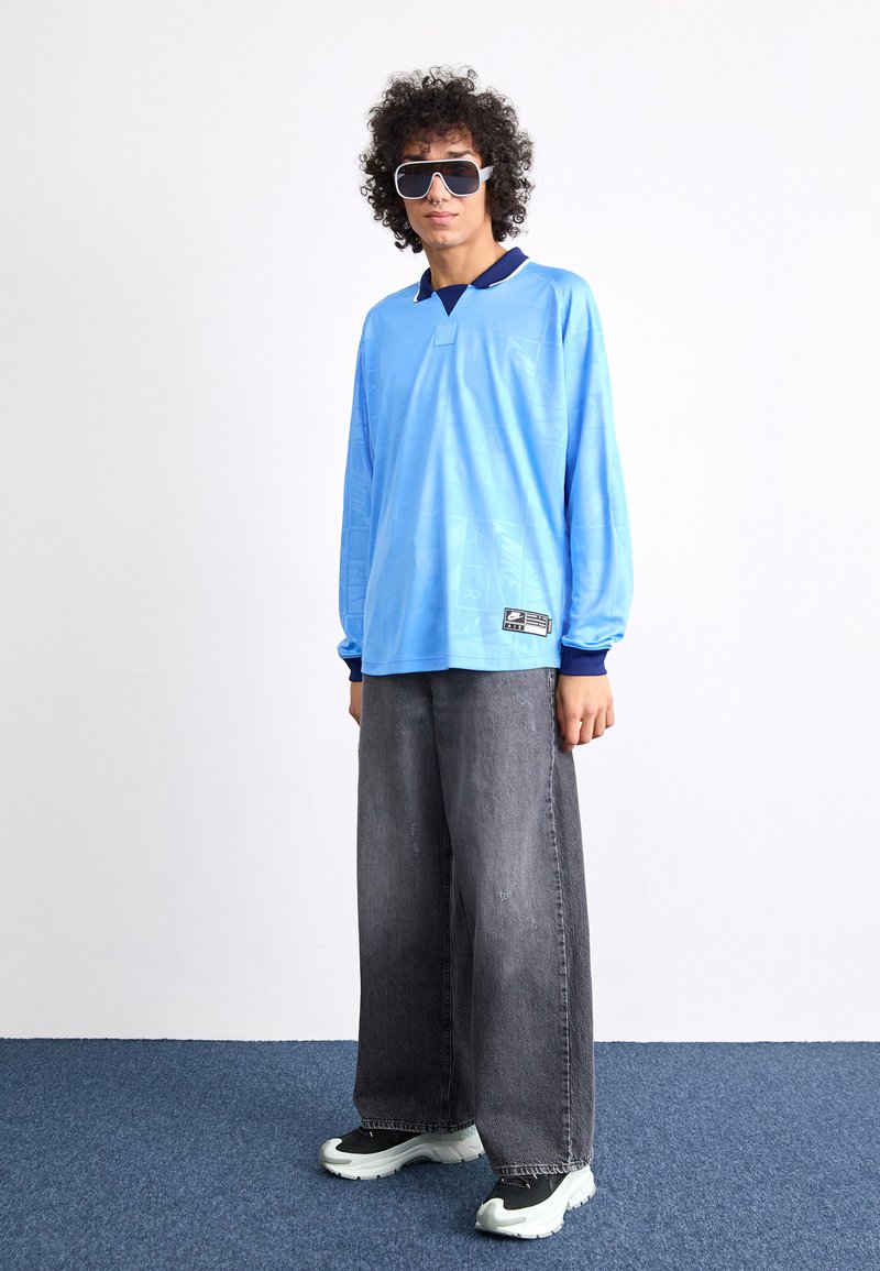 Jasnoniebieska koszula z długim rękawem z granatowym kołnierzykiem, połączona z luźnymi szarymi dżinsami i czarnymi butami sportowymi z białymi podeszwami. Duże, ciemne okulary przeciwsłoneczne.