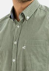 Camisa a cuadros en verde y blanco con cuello abotonado, que presenta botones blancos y un pequeño bolsillo en el pecho. Acento de denim en el cuello.
