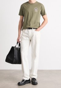 Olivgrön t-shirt med en logotyp, vita byxor, svarta läderskor och en strukturerad svart tygkasse, hålls med ena handen mot en neutral bakgrund.