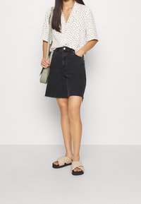 Blusa blanca de manga corta con lunares negros, combinada con una falda de mezclilla negra y sandalias de tira en color beige. Bolsa verde sostenida en la mano.