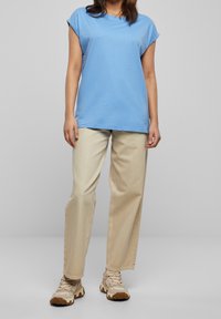 Kvinna som bär en ljusblå kortärmad skjorta, beige raka byxor och beige klumpiga sneakers som står framför en grå bakgrund.