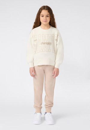Niña pequeña con cabello rizado y largo, vistiendo pantalones de chándal beige, sudadera crema de Champion y zapatillas blancas, de pie contra un fondo simple.